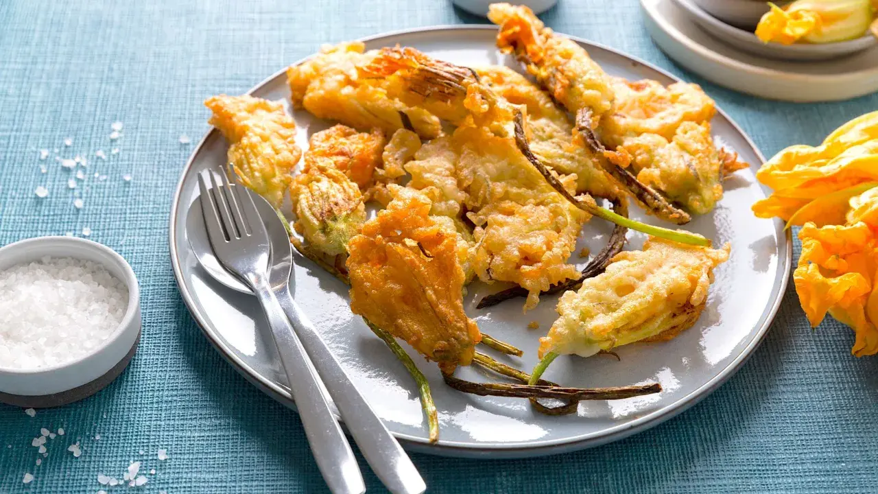 Piatto di fiori di zucca in pastella dorati e croccanti, serviti con sale grosso e posate.