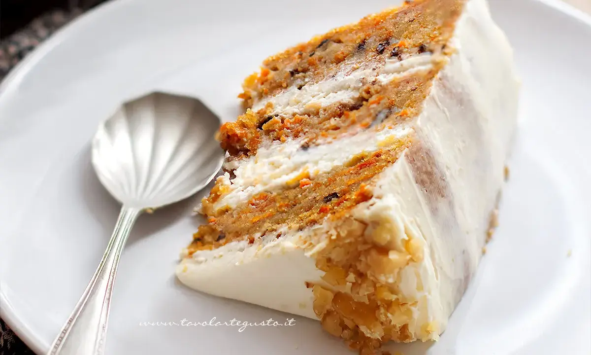 Una fetta di torta di carote e yogurt, soffice e profumata, con glassa bianca e noci tritate.