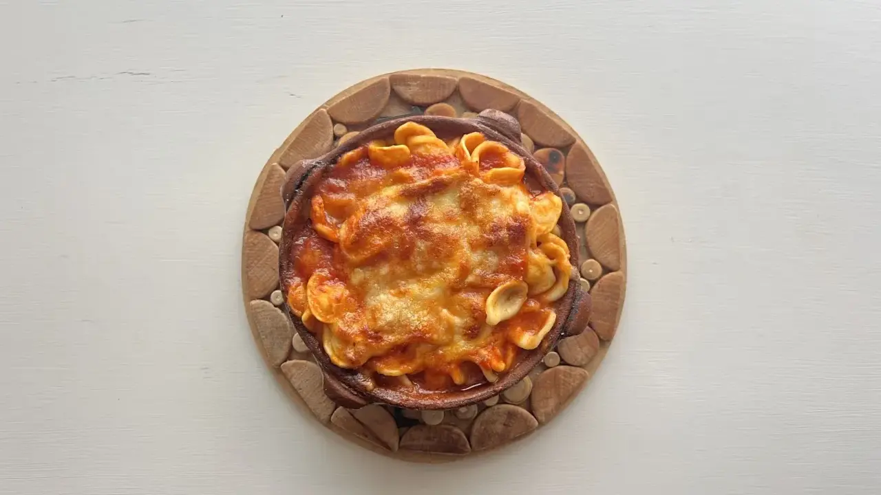 Orecchiette al forno in terracotta, con sugo rosso e formaggio filante, servite su un tagliere di legno.