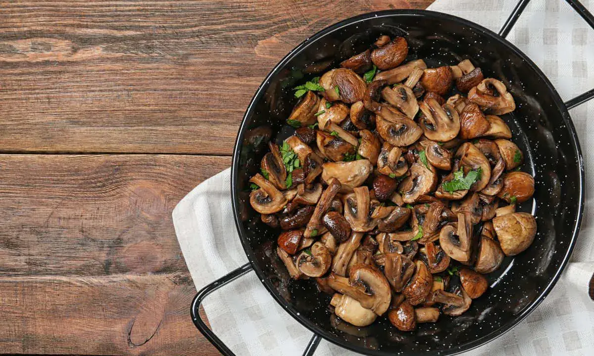 A black pan filled with perfectly cooked, sliced mushrooms and garnished with parsley, sits on a wooden table.