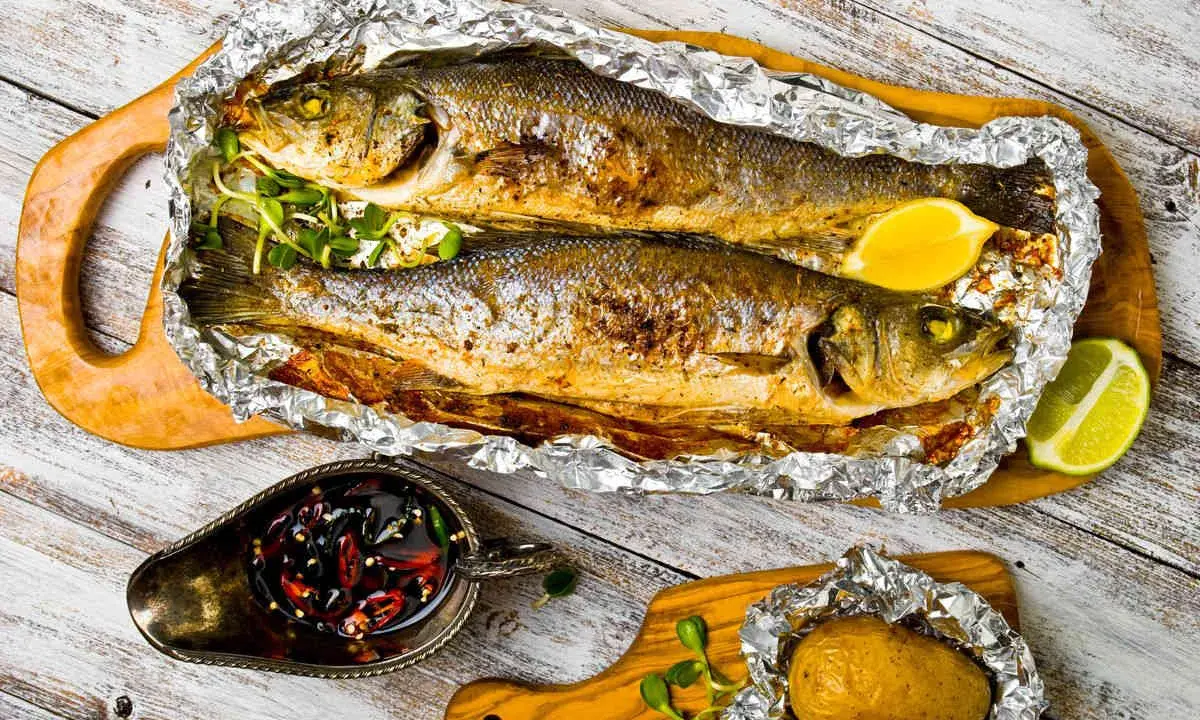 Due branzini cotti al cartoccio, dorati e profumati, serviti con limone, patata al cartoccio e salsa piccante.