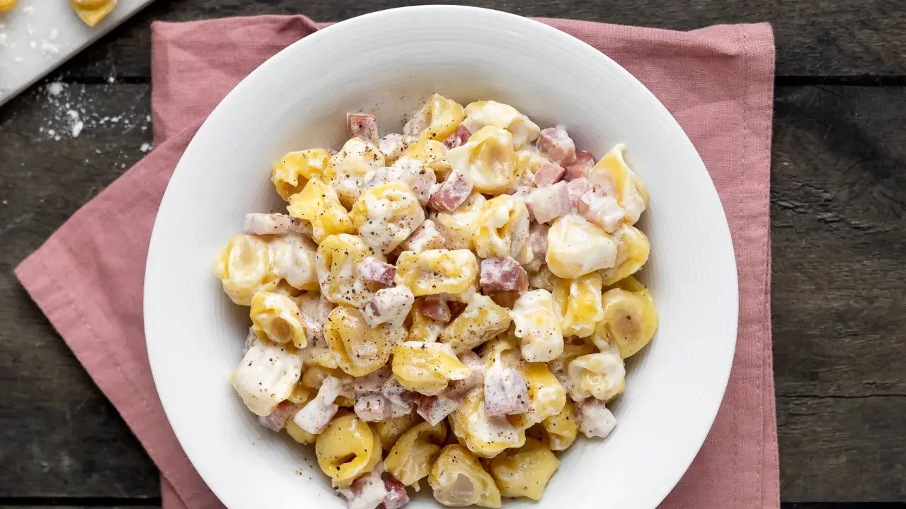 Un piatto invitante di tortellini panna e prosciutto, conditi con pepe nero, su un tovagliolo rosa.