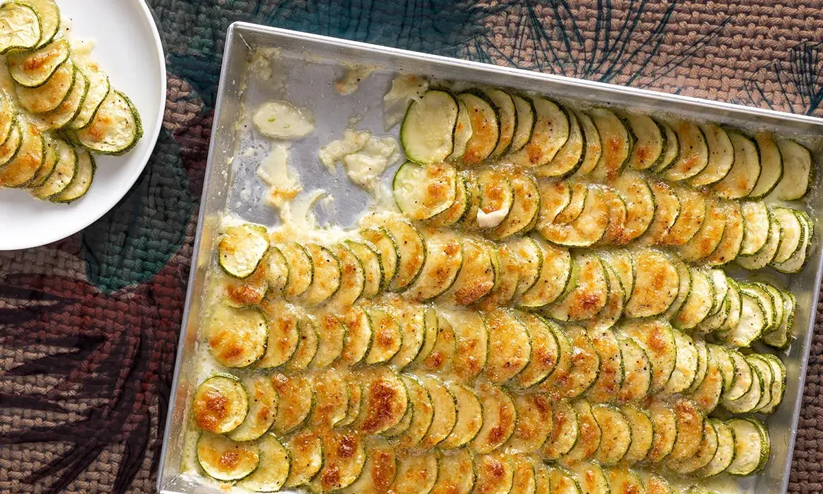 Fette di zucchine al forno con mozzarella filante, disposte in file ordinate in una teglia e su un piatto bianco.