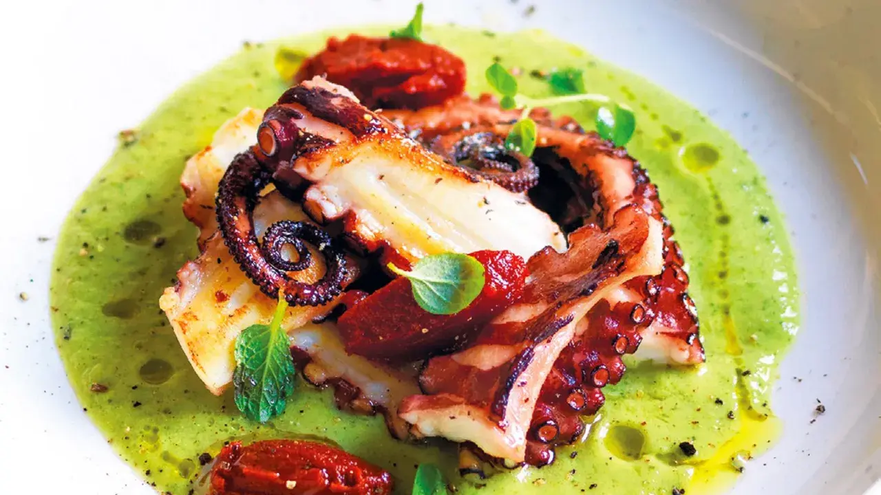 Delizioso polpo alla griglia servito su una crema verde brillante, guarnito con pomodori secchi e erbe fresche.