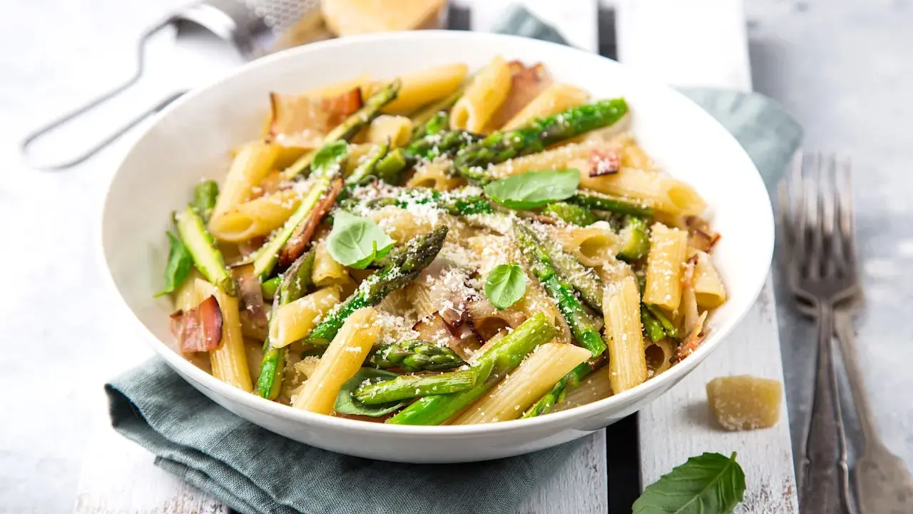 Piatto di pasta con asparagi e pancetta croccante, guarnito con basilico fresco e parmigiano grattugiato.