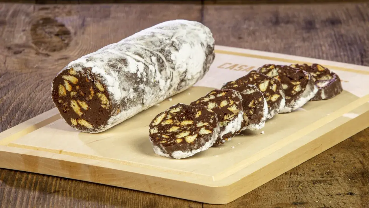 Un rotolo di salame al cioccolato, cosparso di zucchero a velo, affiancato da fette tagliate su un tagliere di legno.
