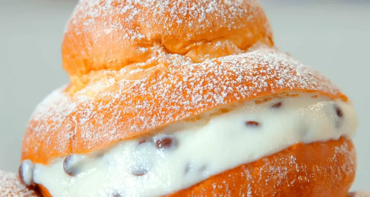 Brioche col tuppo soffice e dorata, ripiena di crema vellutata con gocce di cioccolato, spolverata di zucchero a velo.