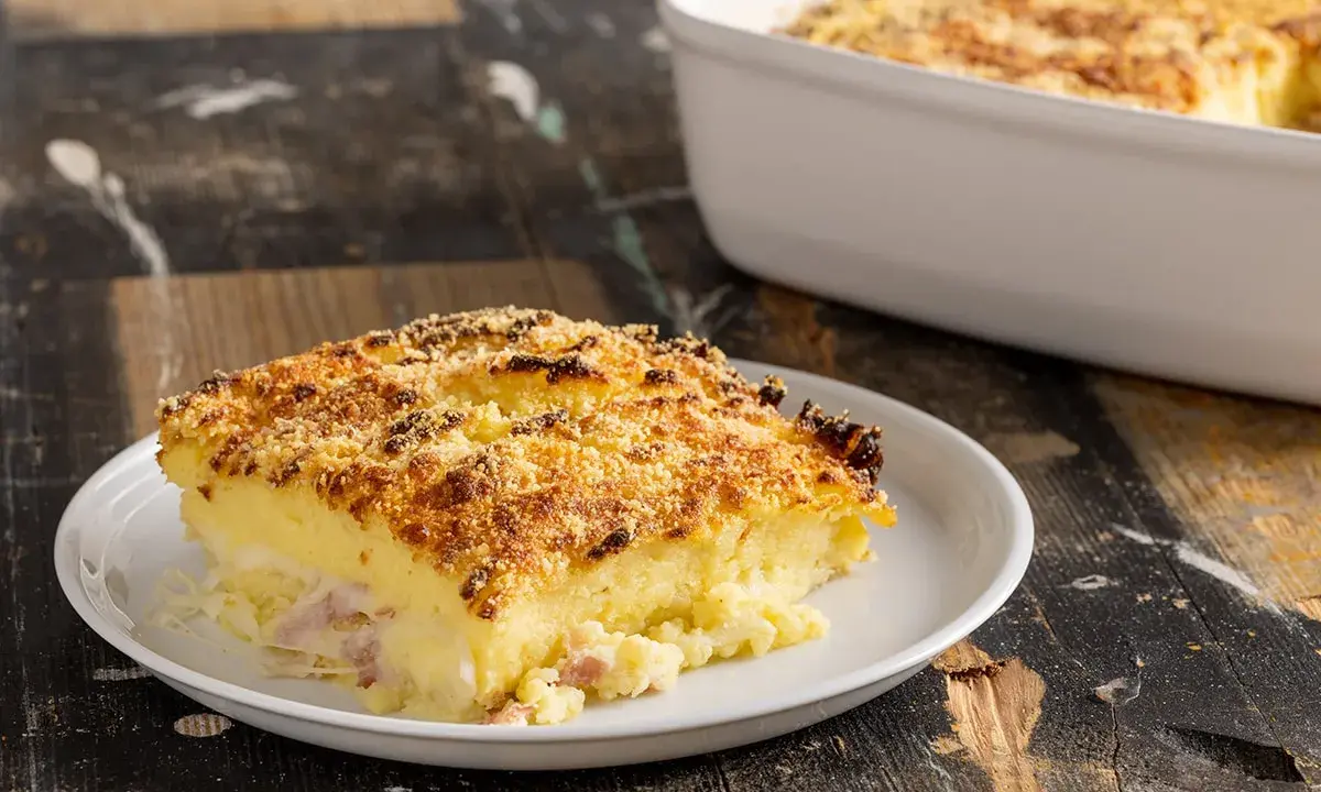 Una fetta di sformato di patate dorato e croccante, con prosciutto e formaggio filante, servita su un piatto bianco.