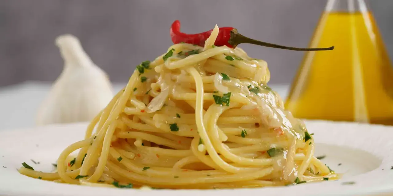 Spaghetti conditi con aglio, olio e peperoncino, guarniti con prezzemolo e un peperoncino rosso. Sullo sfondo, un bulbo d'aglio e una bottiglia d'olio.
