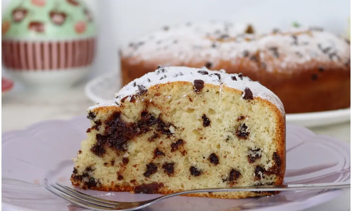 Una fetta di torta soffice con tante gocce di cioccolato, spolverata di zucchero a velo, su un piatto lilla con una forchetta.