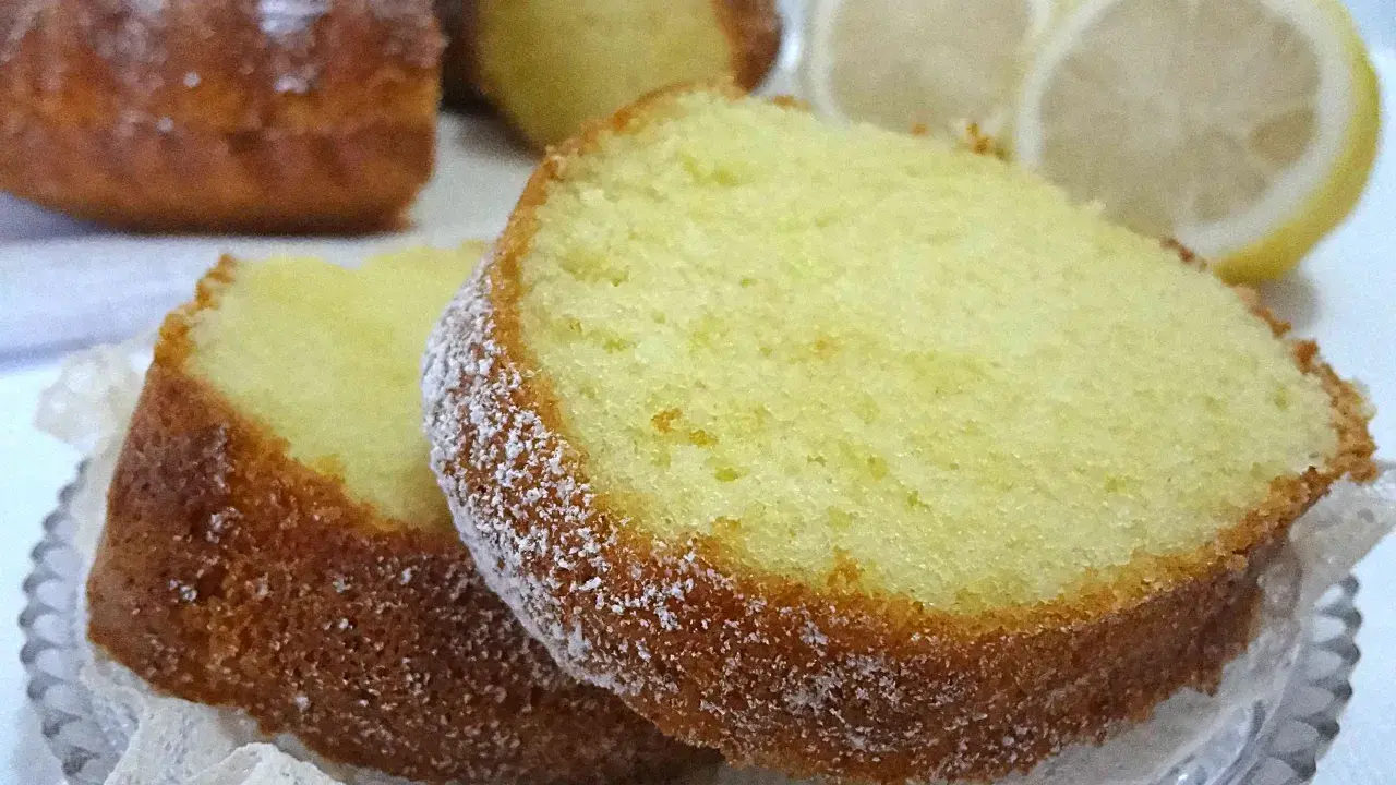 Fette di un soffice ciambellone al limone, spolverate di zucchero a velo, con spicchi di limone sullo sfondo.