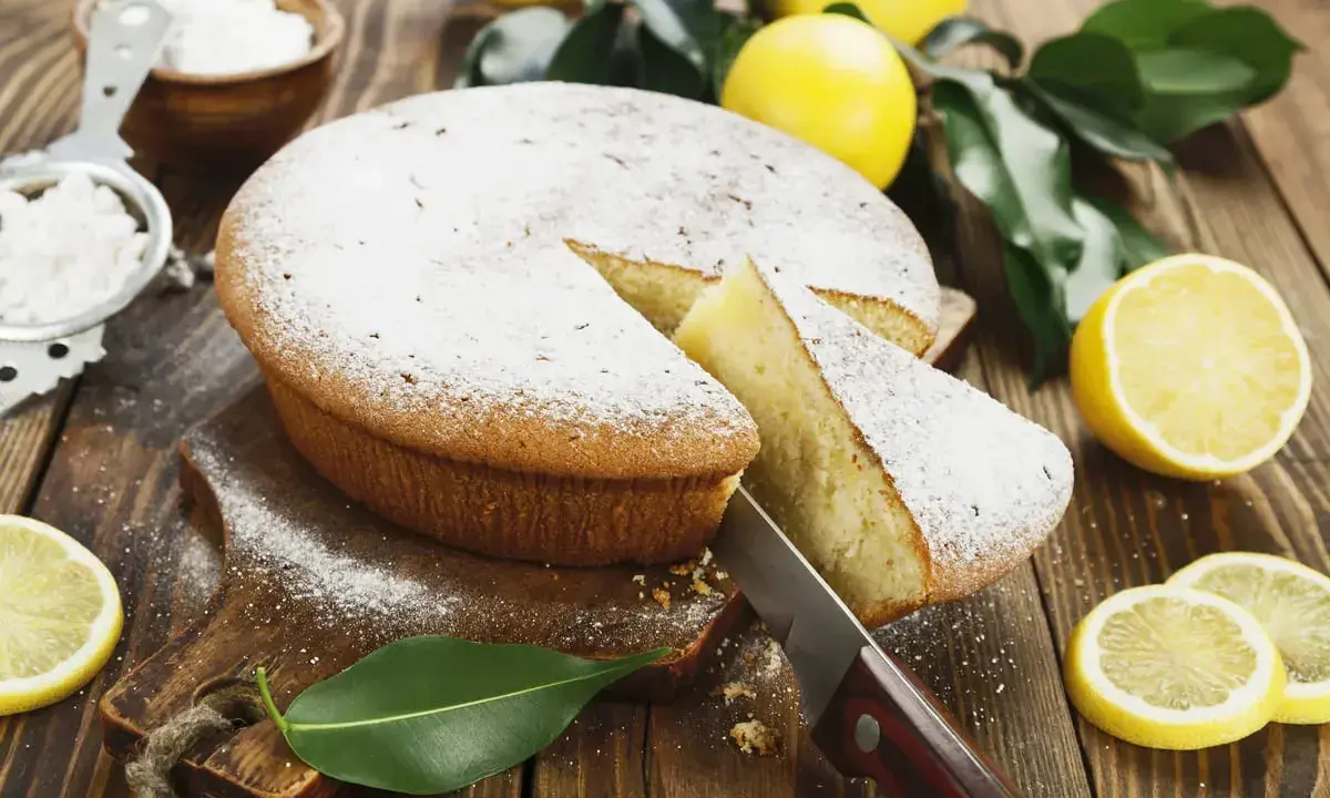Torta al limone velocissima, spolverata di zucchero a velo, con una fetta tagliata e limoni freschi intorno.