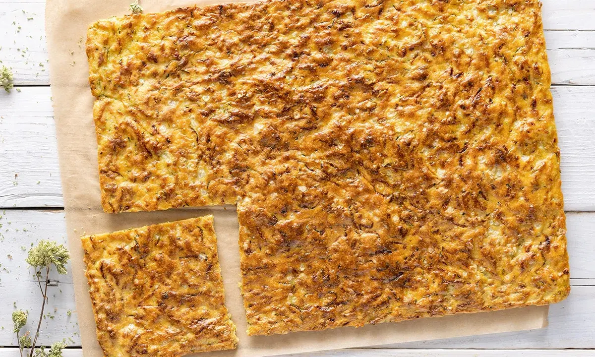 Schiacciata di zucchine dorata e fragrante, tagliata a pezzi su carta da forno, con erbe aromatiche sparse.