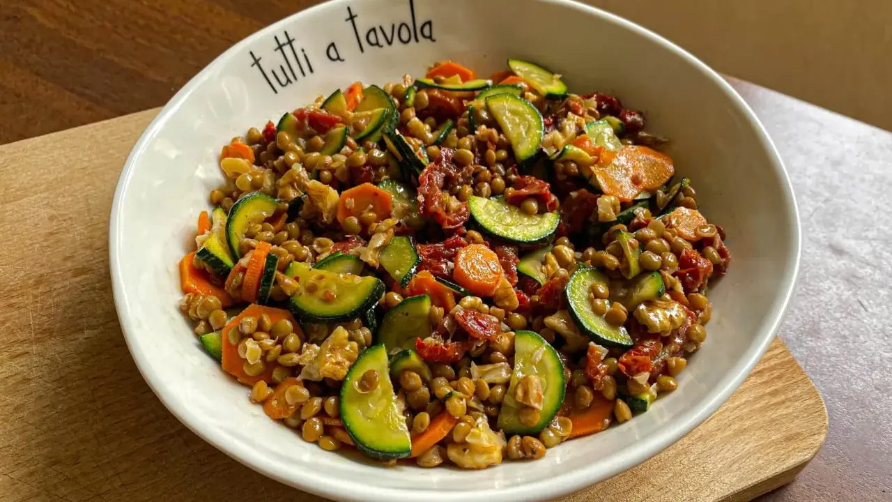 Insalata di lenticchie colorata con zucchine, carote e pomodori secchi, servita in una ciotola con la scritta "tutti a tavola".