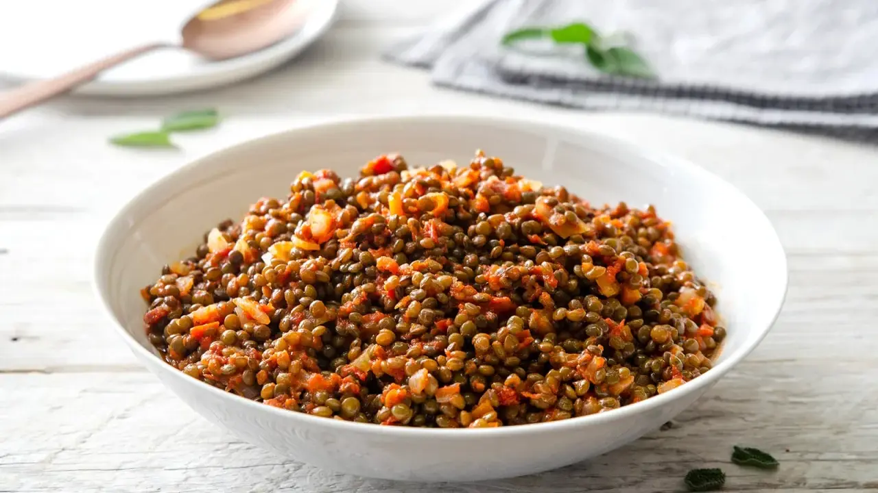 Un piatto invitante di lenticchie in umido, condite con pomodoro e cipolla, servite in una ciotola bianca su un tavolo di legno.