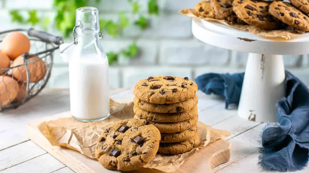 Una pila di biscotti con gocce di cioccolato su un tagliere, accanto a una bottiglia di latte e un cestino di uova.