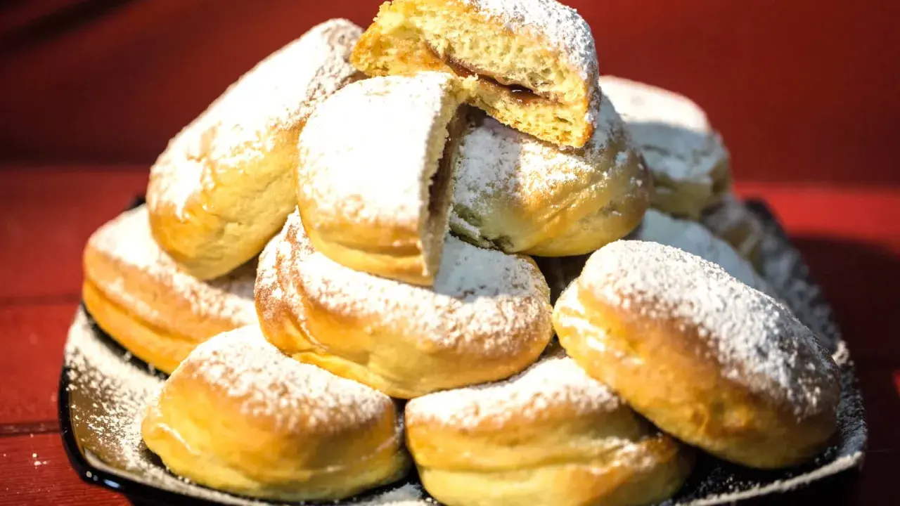 Pile di bomboloni ricetta originale, soffici e cosparsi di zucchero a velo. Uno è tagliato a metà, rivelando un ripieno goloso.