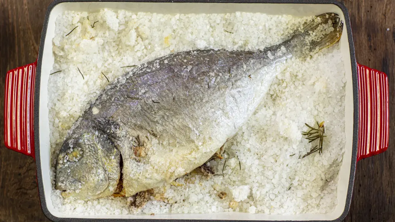 Orata al sale, intera e dorata, adagiata su un letto di sale grosso e rosmarino in una teglia rossa.