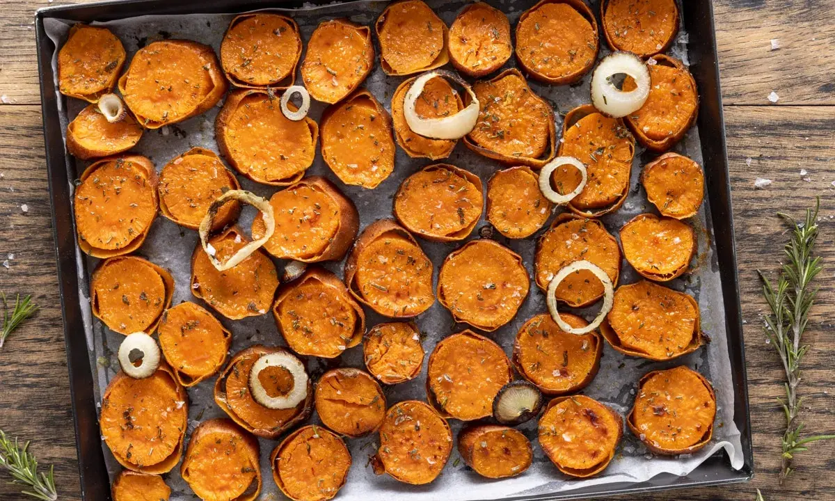 Fette di patate dolci al forno, dorate e condite con erbe aromatiche e sale, disposte su una teglia con anelli di cipolla.