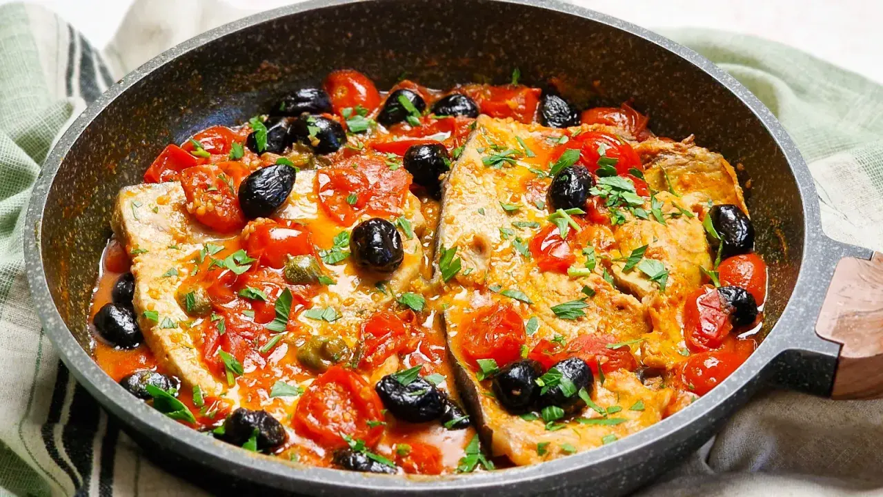 Pesce spada alla siciliana in padella con pomodorini, olive nere e capperi, guarnito con prezzemolo fresco.