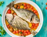 Orata all'acqua pazza, due pesci interi cotti in un brodo saporito con pomodorini e erbe aromatiche, serviti in un piatto bianco su un tavolo di legno blu.