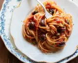 Un piatto di spaghetti alla puttanesca, con pomodoro, olive nere e capperi, servito su un elegante piatto bianco con decorazioni blu.