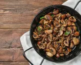 A black pan filled with perfectly cooked, sliced mushrooms and garnished with parsley, sits on a wooden table.