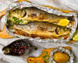 Due branzini cotti al cartoccio, dorati e profumati, serviti con limone, patata al cartoccio e salsa piccante.