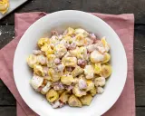 Un piatto invitante di tortellini panna e prosciutto, conditi con pepe nero, su un tovagliolo rosa.