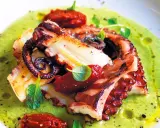 Delizioso polpo alla griglia servito su una crema verde brillante, guarnito con pomodori secchi e erbe fresche.
