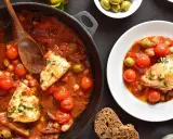 Merluzzo in umido con pomodorini, olive e ceci, servito in una padella e in un piatto, con pane scuro.