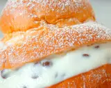 Brioche col tuppo soffice e dorata, ripiena di crema vellutata con gocce di cioccolato, spolverata di zucchero a velo.