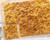 Schiacciata di zucchine dorata e fragrante, tagliata a pezzi su carta da forno, con erbe aromatiche sparse.