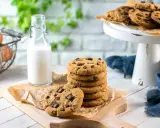 Una pila di biscotti con gocce di cioccolato su un tagliere, accanto a una bottiglia di latte e un cestino di uova.