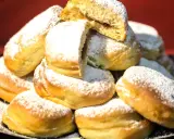 Pile di bomboloni ricetta originale, soffici e cosparsi di zucchero a velo. Uno è tagliato a metà, rivelando un ripieno goloso.