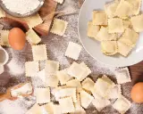 Tanti ravioli fatti in casa sparsi su un tavolo di legno, con farina, uova e una paletta.