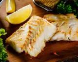 Spigola in padella al vino bianco, dorata e succulenta, servita con limone e insalata fresca su tagliere di legno.
