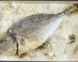 Orata al sale, intera e dorata, adagiata su un letto di sale grosso e rosmarino in una teglia rossa.