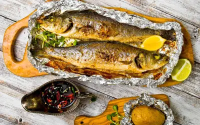 Due branzini cotti al cartoccio, dorati e profumati, serviti con limone, patata al cartoccio e salsa piccante.