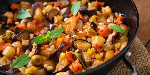 Melanzane in padella, un contorno colorato con carote, olive e pinoli, guarnito con foglie di basilico fresco.