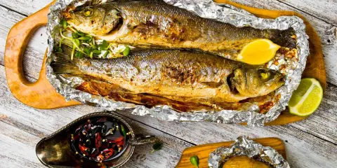 Due branzini cotti al cartoccio, dorati e profumati, serviti con limone, patata al cartoccio e salsa piccante.