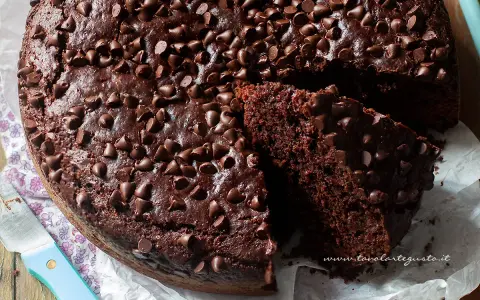 A rich, moist chocolate cake topped with chocolate chips, with a slice cut out to show its texture.