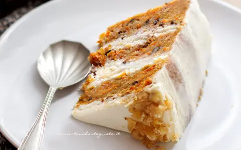 Una fetta di torta di carote e yogurt, soffice e profumata, con glassa bianca e noci tritate.