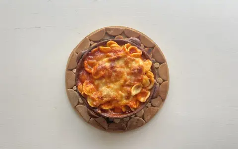 Orecchiette al forno in terracotta, con sugo rosso e formaggio filante, servite su un tagliere di legno.