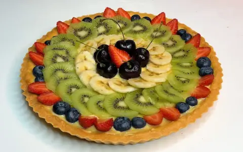 A fruit tart with kiwi, banana, strawberries, blueberries, and cherries on a custard filling.