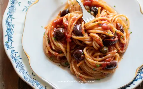 Un piatto di spaghetti alla puttanesca, con pomodoro, olive nere e capperi, servito su un elegante piatto bianco con decorazioni blu.
