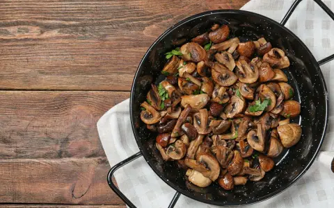 A black pan filled with perfectly cooked, sliced mushrooms and garnished with parsley, sits on a wooden table.