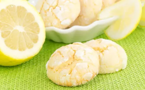Biscotti al limone spolverati di zucchero a velo, accanto a fette di limone fresco su sfondo verde.
