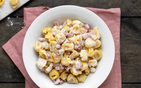 Un piatto invitante di tortellini panna e prosciutto, conditi con pepe nero, su un tovagliolo rosa.