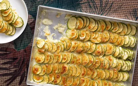 Fette di zucchine al forno con mozzarella filante, disposte in file ordinate in una teglia e su un piatto bianco.